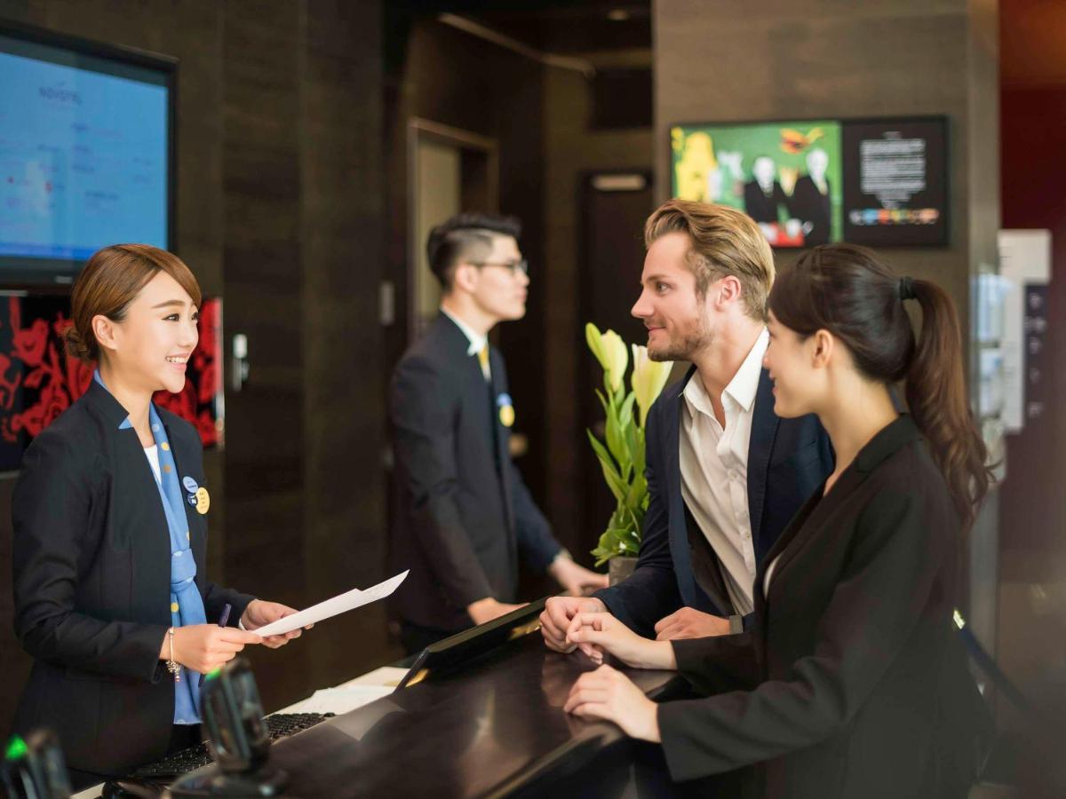 Hyatt Regency Taoyuan International Airport Hotel Exterior photo The photo shows a hotel reception area. There are three staff members behind the reception desk, all dressed in professional attire. One staff member, a woman, is smiling and speaking to two guests—one man and one woman—who are standing at the desk. 