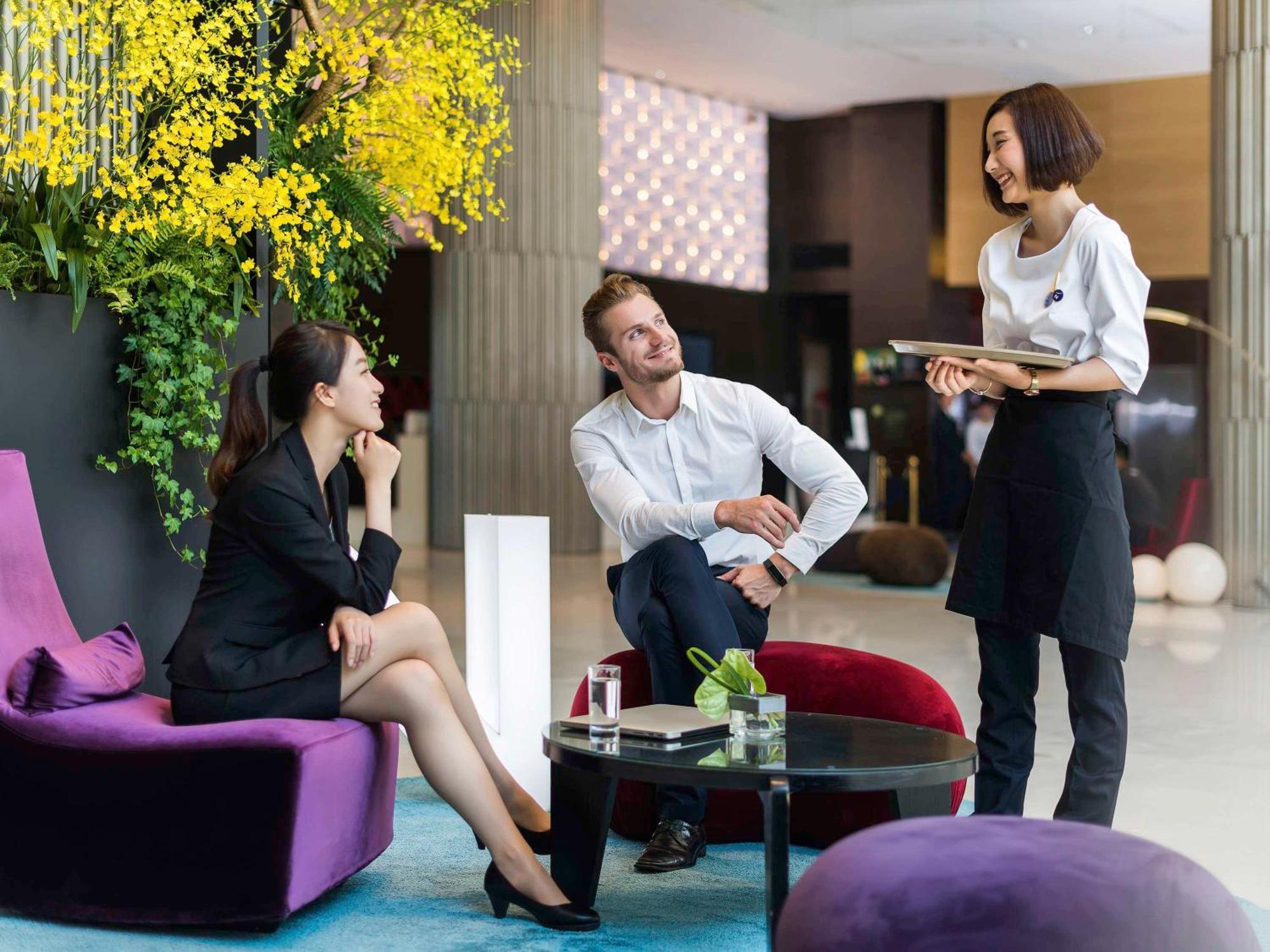 Hyatt Regency Taoyuan International Airport Hotel Exterior photo The photo depicts a casual meeting in a modern, stylish setting. There are three people: two seated and one standing. 

- The woman on the left is dressed in a black suit and appears engaged in conversation. 
- The man in the middle, wearing a light-