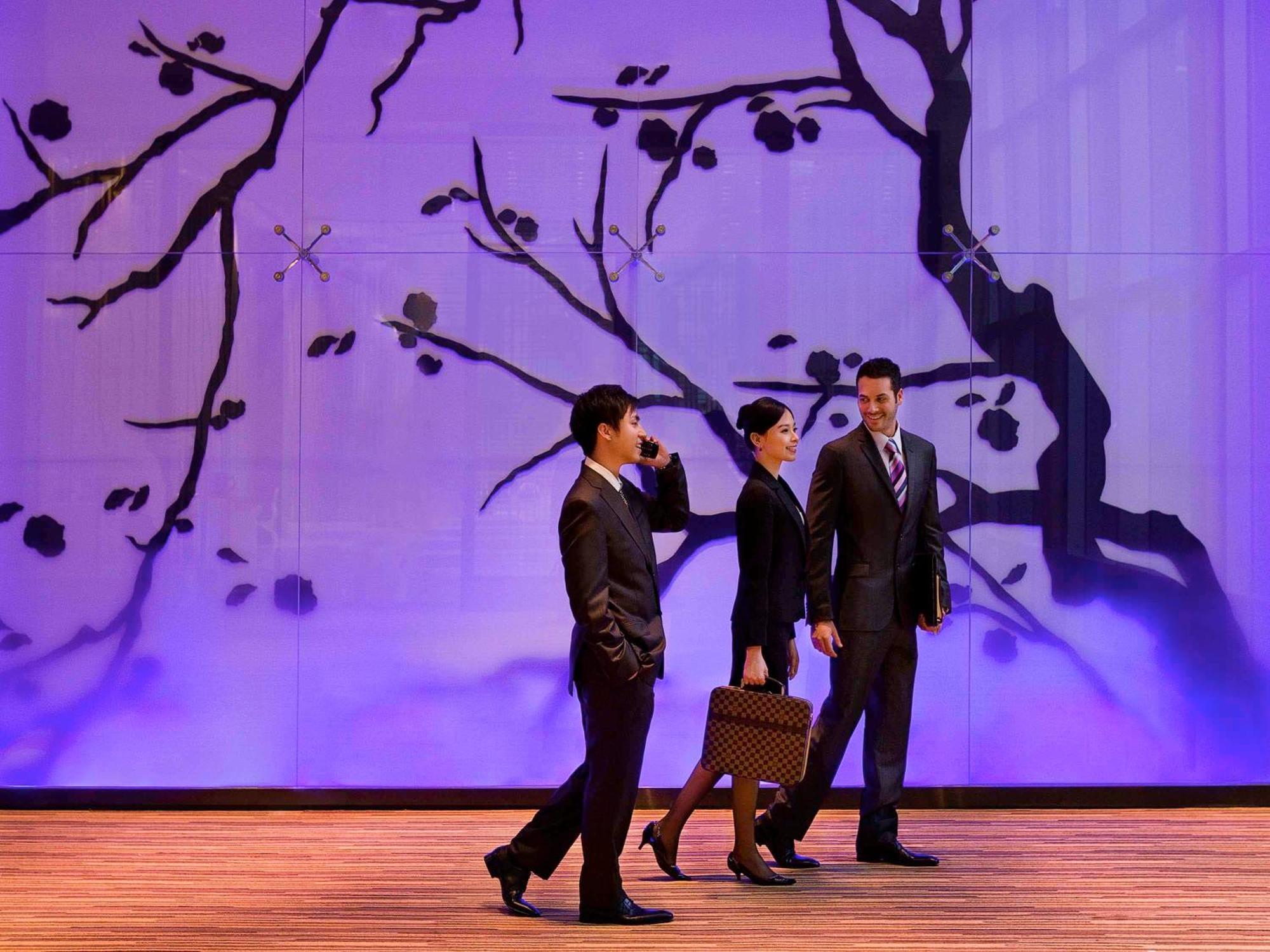 Hyatt Regency Taoyuan International Airport Hotel Exterior photo The photo depicts three professionals walking in a modern, stylish environment. They are dressed in formal business attire—two men in suits and a woman in a dark suit. The background features a large wall with an artistic design of branches, possibly