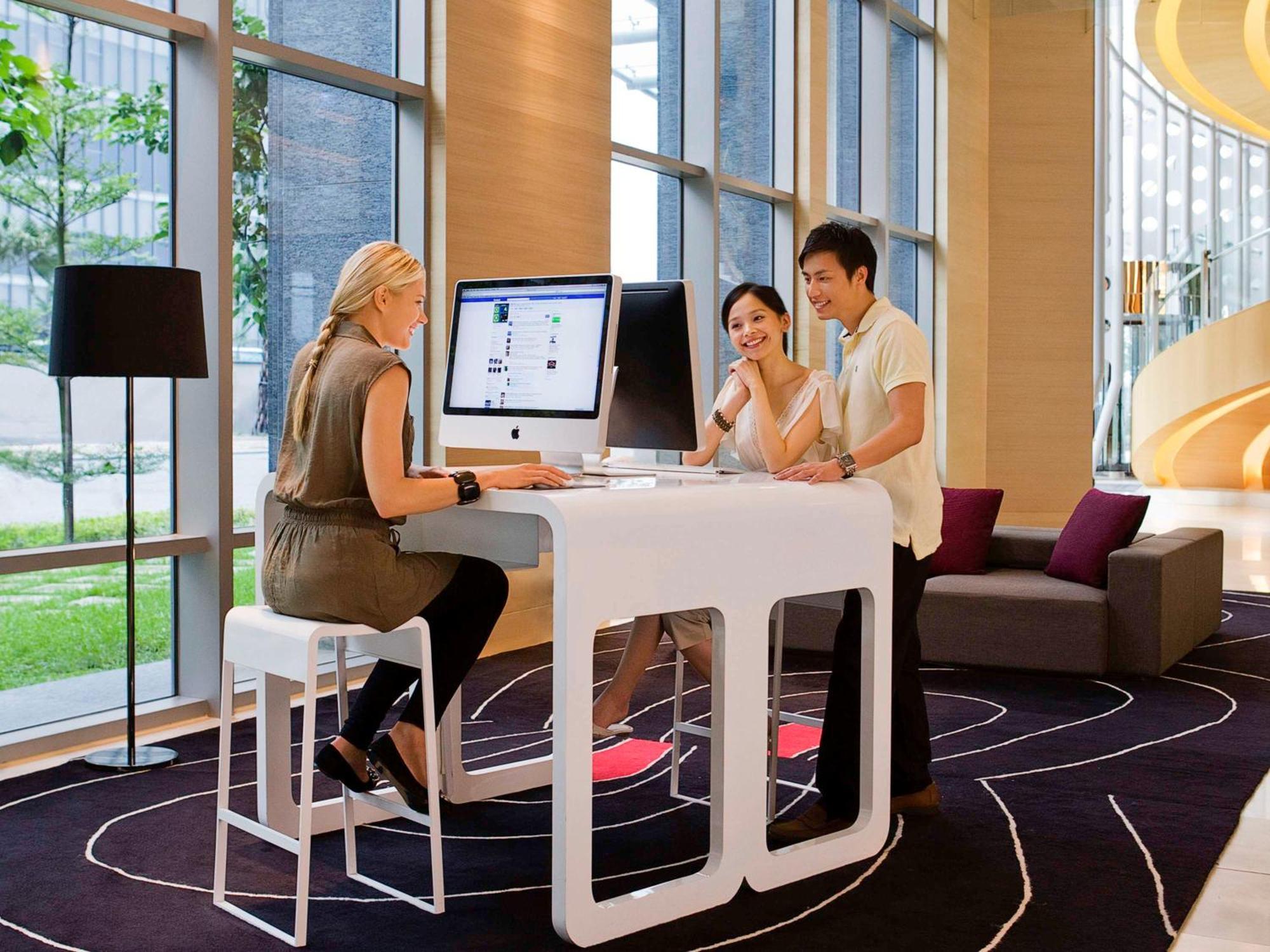 Hyatt Regency Taoyuan International Airport Hotel Exterior photo The photo depicts a modern, well-lit workspace or lounge area. In the scene, there are three individuals engaged in conversation. One person, a woman with long blonde hair, is seated on a high stool in front of a sleek, white desk that holds a comput