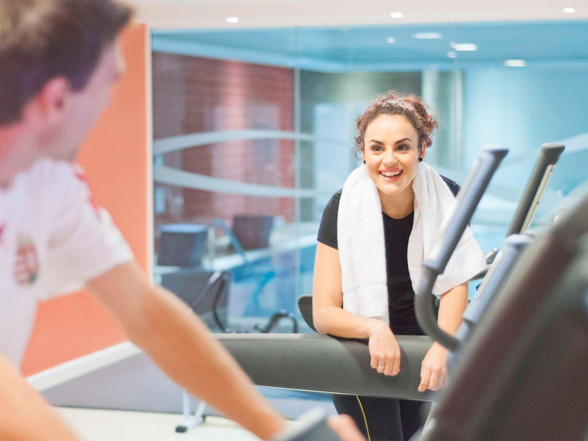 Hyatt Regency Taoyuan International Airport Hotel Exterior photo The photo shows two people in a fitness or gym setting. One person is exercising on an elliptical machine, while the other, wearing a black shirt and a white towel around their neck, is standing nearby and smiling. The atmosphere appears lively and e