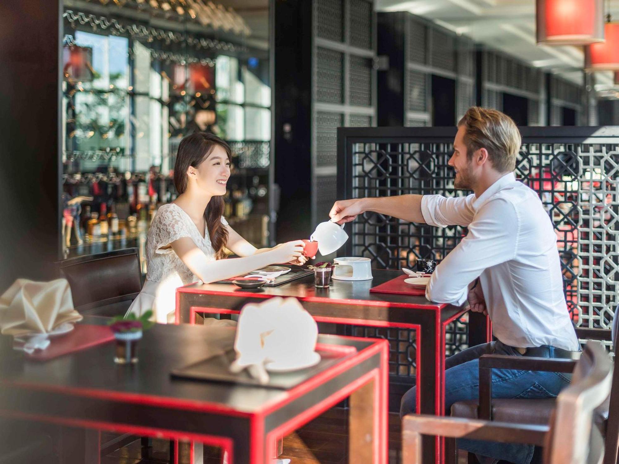 Hyatt Regency Taoyuan International Airport Hotel Exterior photo The photo shows a couple sitting at a table in a restaurant. The woman, wearing a white dress, is smiling at the man, who is dressed in a white shirt. He is pouring something from a teapot or a similar vessel into a cup in front of her. The table is 